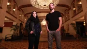 Hosts Katrina Weidman and Nick Groff stand in grand lobby of Statler City serious faces.