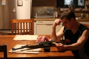 Derek with gun at table, hand on head.