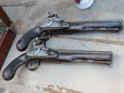 Two flintlock pistols, made of ornate wood and metal, sitting beside each other, ready to be tested.