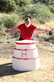 Larry Harley standing behind his massive birthday cake on the shooting range before it is blown to bits with a large "H" printed on it.