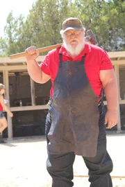 Larry Harley with a huge mallet over his shoulder and leather blacksmith's apron on, poses with the shooting range behind him.