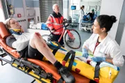 Der Rennrad-Sportler Andreas Lott (Felix Kruttke, l. mit Komparse, h.) wird mit einem offenen Bruch in die Notaufnahme eingeliefert. Dr. Leyla Sherbaz (Sanam Afrashteh, r.) beginnt mit der Erstuntersuchung.