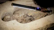 Professor Salima Ikram beleuchtet mit einem Lichtstab eine Mumie in einer 2 600 Jahre alten Grabanlage. Man kann bemalte Kartonnagen und Dutzende von Perlen erkennen, was darauf hindeutet, dass es sich um eine hochkarätige Bestattung handelt.