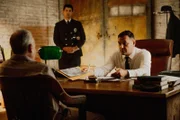 Der Häftling und der Inspektor sitzen am Tisch, während der Polizist steht.