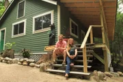Builder Chase and designer Ashley are both sitting on the porch steps together posing for the camera, in the Bullpen cabin build, at Clearwater Lake, Maine, as seen on DIY Network's Maine Cabin Masters.
