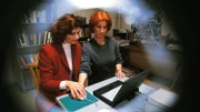 Jeanette beobachtet durch den Türspion, wie Evelyn (Franziska Matthus, li.) Regine (Ana Fonell) eine Computereinführung gibt und sieht auch, wo Regine ihr Manuskript versteckt hat.