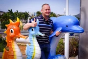 Reiseleiter Bernd posiert am Yalong Bay in Sanya, Hainan, China, mit Seepferdchen und Delfin aus Plastik. Der Strand gilt als einer der schönsten der Welt, doch Sanya ist eine neu entstandene Stadt. Durch den Massentourismus und die kulturellen Unterschiede zu den Chinesen empfindet Bernd viele Dinge als skurril.