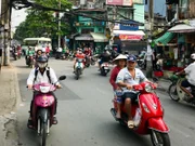 Straßenszene mit zahlreichen Rollern auf der Straße, Läden am Straßenrand und Menschen mit Mundschutz und traditioneller Kopfbedeckung in Ho-Chi-Minh-Stadt (ehemals Saigon), Vietnam.