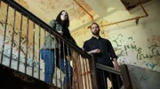 Hosts Katrina Weidman and Nick Groff stand on stairway landing above with decrepit walls behind them.