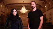 Hosts Katrina Weidman and Nick Groff stand in grand lobby of Statler City Hotel with serious faces.