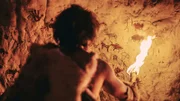 The Riddle of the Carpathian Sphinx, Die Sphinx der Karpaten Primeval Caveman Wearing Animal Skin Standing in His Cave At Night, Holding Torch with Fire Looking at Drawings on the Walls at Night. Cave Art with Petroglyphs, Rock Paintings. Back View