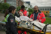 Dr. Lee Evans (Tino Führer, r.) gibt Isa (Lilli Hollunder, l.) Informationen zum Zustand seiner Patientin (Eva Marlen Hirschburger, M.) Die junge Frau wurde angeschossen. (Komparse im Bild)