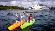 Reiseleiter Moritz und Entertainment Managerin Maika heben, bekleidet mit Schwimmwesten und Kappen, beim Kayak-Fahren vor der Oper und Harbour Bridge in Sydney freudig ihre Paddel in die Luft.