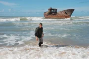 Bear Grylls on beach with shipwreck in background.