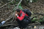 Bear Grylls crouched by stream and eats trout.