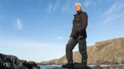 Extreme angler, Jeremy Wade atop rocky terrain, looking out over dramatic coastline.
