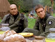 SG-1 wurde von den Enkaranern zu einem Dankesessen eingeladen. Sie sind sehr glücklich über den Planeten, den ihnen das Team ausgesucht hat. Teal'c (Christopher Judge, li.) und Jackson (Michael Shanks) lauschen der Ansprache von Hedrazar.