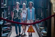 Imogen (Elizabeth Cullen, von links), Lily (Mia Milnes) und Ruksy (Rainbow Wedell) stehen in der Bibliothek der Magie vor einem abgesperrten, verbotenen Gang. Dort hat Professor Maxwell ein Buch versteckt, in das sie unbedingt hineinschauen wollen.