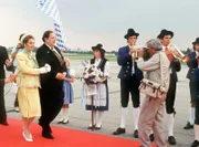 Standesgemäß wird die Königin von Mandalia (Michaela May, l) vom Ministerpräsidenten (Georg Marischka) auf dem Flughafen in Empfang genommen. Herbie (Dieter Hildebrandt, r) ist dabei voll in Aktion.