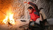 Bear Grylls lays in front of burning fire in man-made cave.