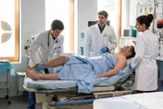 Ben Ahlbeck (Philipp Danne, l.) untersucht unter den gestrengen Augen von Dr. Niklas Ahrend (Roy Peter Link, M.) und Dr. Leyla Sherbaz (Sanam Afrashteh, r.) Marcel Witte (Martin Becker, liegend) nach seinem Autounfall auf Verletzungen an der Hüfte.