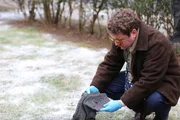 Reconstruction Actor playing Det Schnell (Scott Watson) in brown jacket and yellow glasses crouching to investigate a piece of clothing