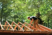 Builder Ryan is standing on the roof during demolition of the cabin roof, for Vince and Nicole's cabin build, in Desert Pond, Maine.