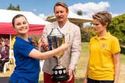 Camilla (Agnes Mohan, l.) nimmt stolz den Pokal aus den Händen von Jared (Paul Bishop, M.) für ihre Schulmannschaft entgegen. Nach einem Einspruch wurde ihnen nachträglich der Sieg zugesprochen. Imogen (Elizabeth Cullen, r.) ist sauer, denn eigentlich hätte ihre Schule den Pokal verdient.
