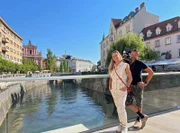 Ljubljana ist mit rund 300.000 Einwohner:innen nicht nur die Hauptstadt, sondern zugleich auch die größte Stadt Sloweniens. Die Universitätsstadt liegt direkt im Herzen Sloweniens und ist nicht nur äußerst grün, sondern auch sehr nachhaltig – 2016 wurde sie als European Green Capital ausgezeichnet. Doch wie lässt sich die Stadt mit schmaler Reisekasse erleben? Reichen 300 Euro für 2 Personen und 2 Tage aus?  In der neuen Folge „2 für300 – Ljubljana“ wollen Moderatorin Tamina Kallert und Kameramann Uwe Irnsinger genau das herausfinden. Bei dem Angebot aus Kultur, Stadt und Natur haben sie wie immer ein Budget von 300 Euro.  (v.l.n.r.) Tamina und Uwe stehen auf einer Brücke in Ljubljana – im Hintergrund die Franziskaner Kirche