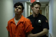 Craig Rabinowitz being led in an orange jumpsuit down a hallway by a police officer.