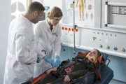 Sandra Schüttler (Anna-Lena Schwing, r.) wird nach einem Sturz mit ihrem Motorrad in die Notaufnahme eingeliefert. Dr. Moreau (Mike Adler, l.) und Elly Winter (Juliane Fisch, M.) untersuchen sie auf mögliche Wirbelverletzungen.