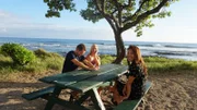 Home buyers Rachael Adair (M), Dan Adair (L) and Realtor Rebecca Morton (R) sitting at a picnic table by the ocean as seen on Hawaii Hunters (Action)