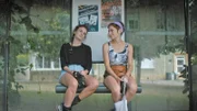Von links: Zoë (Violet Braeckman, l.) und Ylena (Laurian Callebaut, r.) sitzen auf der Bank einer Bushaltestelle. Werden sie ihre Freundin Lisa wiederfinden?