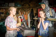 Der maskierte Täter (Luise Wolfram) hat Pinja (Sina Michel) in seiner Gewalt und fordert von Stella (Zoë Malia Moon, links) den Koffer mit den Schmetterlingspuppen zurück.