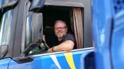 Manuel sitzt im Lastwagen, schaut aus dem Fenster, streckt die Ellbogen aus dem Fenster und lächelt