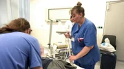 Nurse Karoline is holding some cables in her hand while apparently talking to a blurred patient. In the back we see a young doctor in a white coat, to the right a carry on bagand a chair. A male long haired nurse is seen on the left front.