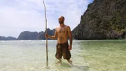 Ed Stafford standing, holding a bamboo rod with the sea and cliffs behind him.