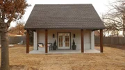 Asymmetrically stylish, the fire pit on the front porch adds interest and begs for attention, whereas the symmetry of the build itself, offers a stately simplicity. The front pillars compliment the leaves on the dormant tree and the roof compliments the picket fence behind, all of which, contrasts the painted brick and faux shake shingle siding, in El Reno, Oklahoma, as seen on Tiny House, Big Living.