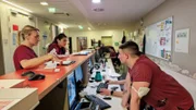 Some members of staff are talking and listening to each other at the orange counter. Y female nurse wearing glasses and a female physician are leaning on, a young male nurse with a mask hanging on his arm is listening.