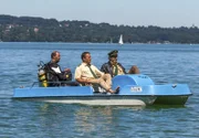 Hubert (Christian Tramitz, Mitte) und Staller (Helmfried von Lüttichau, rechts) sitzen in einem Tretboot und fahren auf den See hinaus. Yazid (Hannes Ringelstetter) sitzt hinter ihnen in Tauchermontur. Er soll für Hubert und Staller testen, ob man mit einem Schlittschuh unter Wasser an einer Boje hängen bleiben kann.