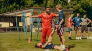 Carlo Brenner (Jeremias Meyer, l.) verletzt sich beim Schuss und liegt am Boden. Während Aydin Alkan (David Ali Rashed, 3.v.l.) sein Bein dehnt, nutzt Trainer Andi (Lukas 'Cossu' Staier, 2.v.l.) die Gelegenheit für eine Ermahnung. Drei Mitspieler (Komparsen) beobachten das Geschehen.