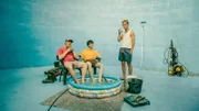 BlaBla (Sebastian Jakob Doppelbauer, l.), Carlo (Jeremias Meyer, M.) und Aydin (David Ali Rashed, r.) haben eine Güllegrube blau gestrichen und wollen daraus ihren eigenen Pool mit Beach machen – bei den heißen Temperaturen in Hintervorderbach ist das die einzige Möglichkeit, sich abzukühlen.