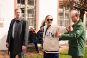 Der „geflüchtete“ Fußballtrainer (Peter Kollmann, M.) ist außer sich - keiner glaubt ihm, dass Riedl ihn angestiftet hat (Stefan Kaminsky als Anstaltsleiter, l. und Komparse, r.).