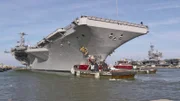 Zwei Spezialschlepper helfen dem 95 tausend Tonnen schweren Flugzeugträger vor Beginn des Manövers aus dem Hafen hinaus auf offene See zu steuern.