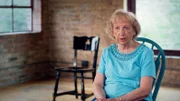 Ida sitzt vor der Kamera. Im Hintergrund ist ein leerer Stuhl zu sehen, er soll die verschollene Schwester Genia symbolisieren.