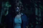 A young man smokes a cigarette in the woods.