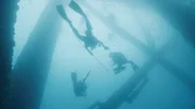 Taucher schwimmen um die Pfähle einer Offshore-Ölplattform, mit Markierungsspeeren in der Hand.