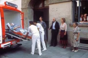 Die Hausbesitzerin Frau Winter (Erika Wackernagel, links) muß ins Krankenhaus. Von links: Ihr Neffe, Herr Eggerer (Gerhard Zemann), der Mieter Herr Schrambeck (Helmut Brasch), die Hausmeisterin Martha Haslbeck (Veronika Fitz), eine Anwohnerin (Komparsin) sowie die Mieter Rosa (Sarah Camp) und Hans Ostermeier (Hans Stadlbauer) beobachten das traurige Ereignis.