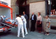 Die Hausbesitzerin Frau Winter (Erika Wackernagel, links) muß ins Krankenhaus. Ihr Neffe, Herr Eggerer (Gerhard Zemann), der Mieter Herr Schrambeck (Helmut Brasch), die Hausmeisterin Martha Haslbeck (Veronika Fitz), eine Anwohnerin (Komparsin) sowie die Mieter Rosa (Sarah Camp) und Hans Ostermeier (Hans Stadlbauer) beobachten das traurige Ereignis.