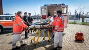 Dr. John Schmidt (Joel Williams, r.) ist beunruhigt wegen der Werte seines Patienten (Leon Blaschke, M.). Es könnte sich um eine Vergiftung handeln. Mattes Seeler (Matthias Schloo, 3.v.l.) und Melanie Hansen Sanna Englund, 2.v.l.) überwachen den Abtransport.